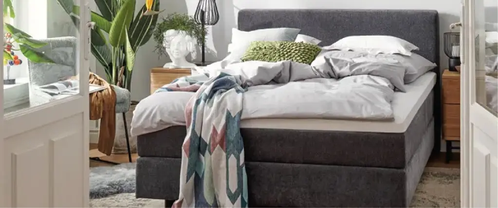 Modern eingerichtetes Schlafzimmer mit hochwertigem Polsterbett in Grau, gemütlicher Bettwäsche, Holznachttischen und Pflanzen. Stilvolle Schlafzimmereinrichtung mit Fokus auf Komfort, Design und wohnlicher Atmosphäre.